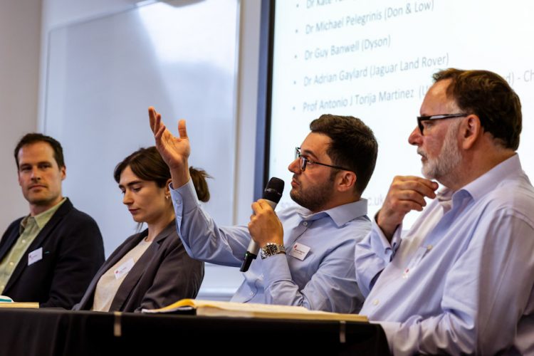 Four panel members sit at a table during a conference discussion. One panelist speaks into a handheld microphone while gesturing toward the audience. A presentation slide with text is projected on a screen behind the panel, and name badges, notebooks and microphones are visible in the meeting room setting.