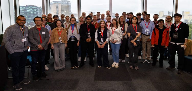 A large group of conference participants wearing red lanyards pose for a group photograph in a bright indoor space. Floor-to-ceiling windows behind the group overlook an urban cityscape with modern buildings. The participants stand in several rows facing the camera, with conference badges visible and natural daylight illuminating the room.