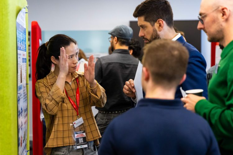 Attendees gather around a research poster during a conference poster session. One participant wearing a conference badge gestures with both hands while explaining the poster to several listeners standing nearby. The display board is positioned against a bright green partition, and other participants and poster presentations can be seen in the background.