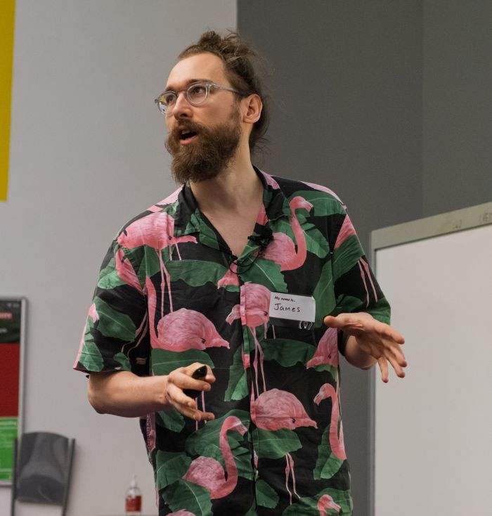 A speaker, James Hipperson, stands indoors delivering a presentation while holding a remote control. The speaker wears glasses and a black shirt with a pink flamingo pattern and a visible name badge. A whiteboard and grey wall are visible in the background, indicating a lecture or conference setting.
