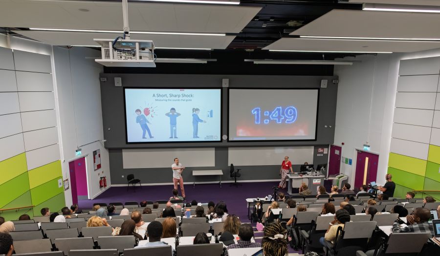 A wide view of a lecture theatre during a presentation event. Audience members are seated in tiered rows facing a stage, where a presenter, Matt Torjussen, stands beneath two large projection screens. One screen displays a slide titled “A Short, Sharp Shock,” while the other shows a countdown timer reading 1:49. Event staff are seated at a desk to one side of the stage, and the lecture theatre features grey walls, purple flooring, and green accent panels.