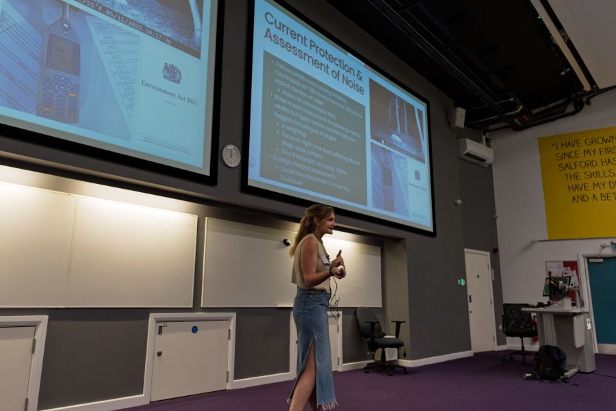 A presenter, Sarah Armstrong, stands at the front of a lecture theatre delivering a talk, with two large projection screens displaying a slide titled “Current Protection & Assessment of Noise.” The slide includes text, photographs, and an image of a sound level meter. Whiteboards line the wall behind the presenter, and a lectern and equipment desk are visible to the side. The room has purple carpeting and auditorium-style presentation facilities.
