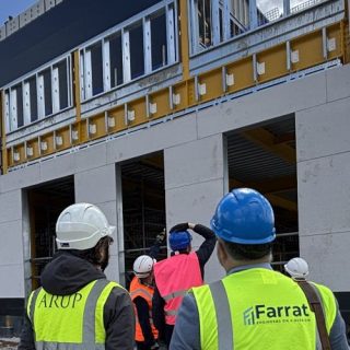 A group of visitors wearing high-visibility safety vests and hard hats stand outside a multi-storey building under construction during a site tour. The building features exposed structural framing, scaffolding and partially completed exterior walls. Several attendees gather in front of the structure while others walk across the gravel construction site. Logos for engineering firms ARUP and Farrat are visible on some safety vests. The scene is set under a bright blue sky with large clouds, highlighting a modern construction and engineering project.