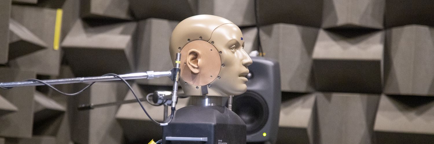 A head-and-torso simulator used for acoustic measurements is positioned inside an anechoic chamber lined with wedge-shaped sound-absorbing foam. Measurement microphones and cables are attached near the artificial ears, while a loudspeaker stands in the background. The specialised equipment is used to study how sound is perceived and recorded in a controlled, reflection-free acoustic environment.