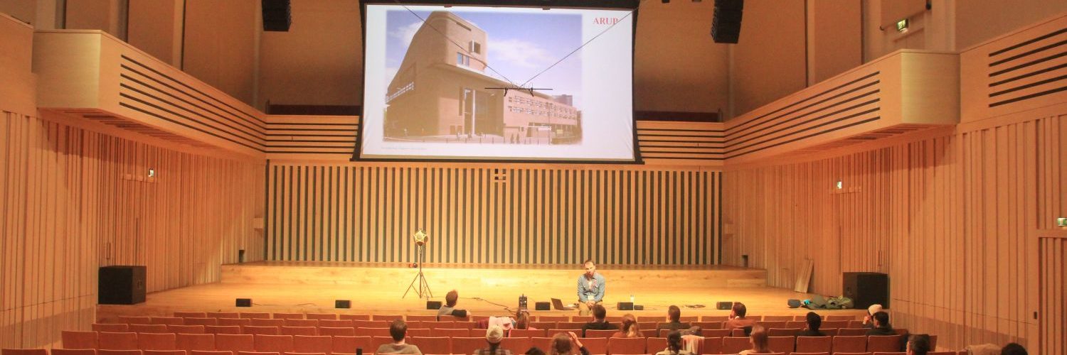 A lecture hall with rows of wooden auditorium seating is being used for an acoustics demonstration or presentation. A small audience is seated facing a stage, where a presenter sits near audio equipment and a microphone setup. A large projection screen displays an image of a building, and acoustic measurement equipment is positioned at the front of the hall. The venue features timber wall panelling and a high ceiling designed to support high-quality sound performance.