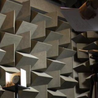An acoustics experiment takes place inside an anechoic chamber lined with large wedge-shaped sound-absorbing panels. A loudspeaker mounted on a stand faces measurement equipment positioned on a tripod, while a researcher stands nearby holding a notebook. The chamber’s geometric foam wedges cover the walls to minimise sound reflections, creating a controlled environment for acoustic testing and measurement.
