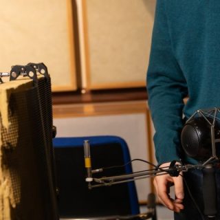 An acoustics research experiment is set up in a laboratory containing sound-absorbing wall panels. A person stands beside a microphone mounted on a stand and a test panel made of acoustic material. Additional specialised equipment, including a head-shaped acoustic measurement device, is visible on a table in the background. The scene shows a controlled environment for studying sound transmission, recording or acoustic performance.