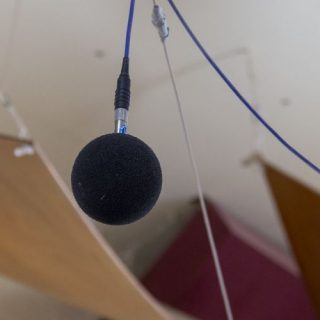 A suspended measurement microphone hangs from cables beneath a ceiling fitted with curved wooden acoustic panels. The microphone is positioned among tensioned wires and acoustic treatment elements designed to control sound within the space. The background is softly out of focus, highlighting the microphone and the architectural acoustics features of the room.