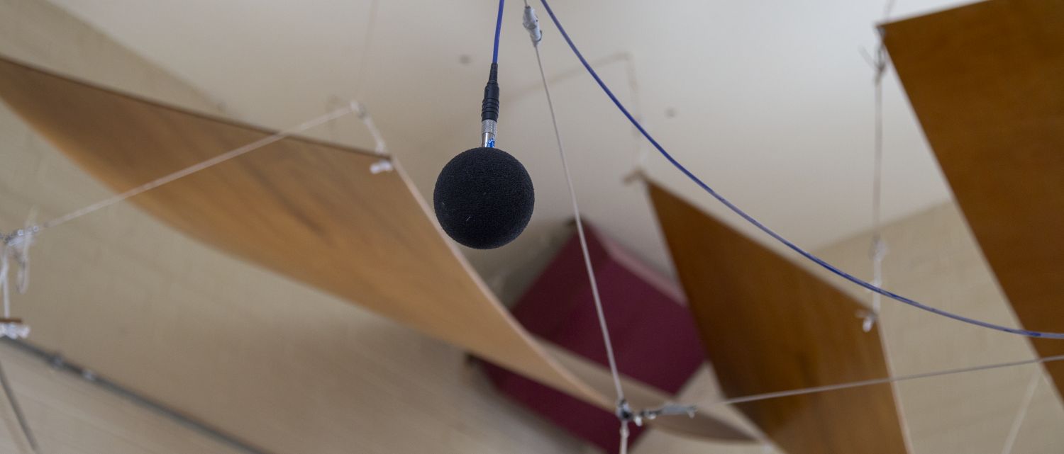 A suspended measurement microphone hangs from cables beneath a ceiling fitted with curved wooden acoustic panels. The microphone is positioned among tensioned wires and acoustic treatment elements designed to control sound within the space. The background is softly out of focus, highlighting the microphone and the architectural acoustics features of the room.