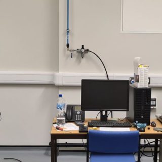 Laboratory testing equipment is arranged inside an acoustics research facility. Two large materials testing machines stand on either side of a desk with a computer workstation, monitor and telephone. A banner for the UK Acoustics Network (UKAN) is displayed on the left, featuring the text “The Focal Point of Acoustics.” Additional laboratory equipment, cables and a blue trolley are visible around the room, highlighting a controlled environment for acoustics and materials research.