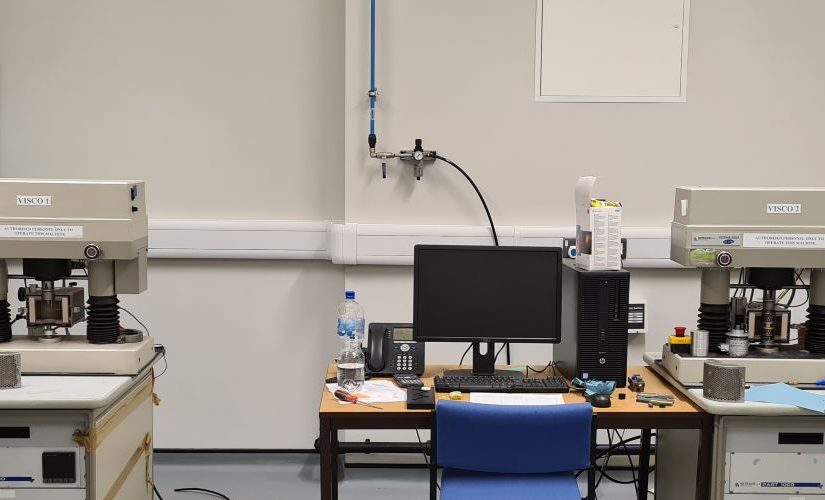 Laboratory testing equipment is arranged inside an acoustics research facility. Two large materials testing machines stand on either side of a desk with a computer workstation, monitor and telephone. A banner for the UK Acoustics Network (UKAN) is displayed on the left, featuring the text “The Focal Point of Acoustics.” Additional laboratory equipment, cables and a blue trolley are visible around the room, highlighting a controlled environment for acoustics and materials research.