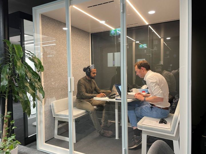 Two men sitting in sound proof booth working on laptops doing a listening test using headphones