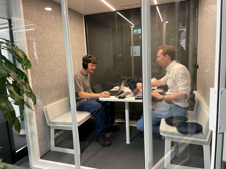 Two men sitting in sound proof booth working on laptops doing a listening test using headphones.