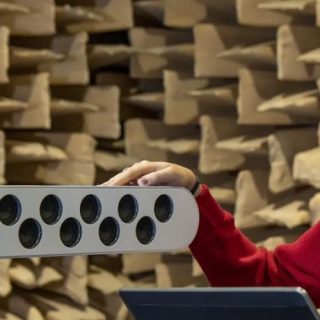 A researcher operates a laptop beside a linear microphone array inside an anechoic chamber lined with wedge-shaped sound-absorbing foam panels. The microphone array contains multiple evenly spaced sensors arranged in a straight line on a stand, while additional acoustic measurement equipment is visible in the foreground. The specialised testing environment is designed to eliminate sound reflections, allowing precise acoustic measurements and sound source localisation research.