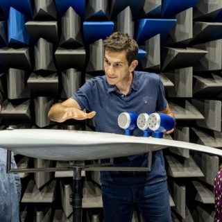 Three people stand around an aircraft model inside an anechoic chamber lined with grey and blue acoustic foam wedges. One person gestures above the model while explaining a concept, and the other two observe. The aircraft model is mounted on a stand and fitted with small cylindrical devices, suggesting an acoustics or aerodynamics experiment in a specialised research environment.