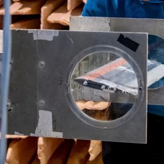 A researcher adjusts a piece of experimental equipment inside an anechoic chamber lined with wedge-shaped sound-absorbing panels. The setup includes a curved acoustic test section with metal and transparent components, allowing observation of a small test model positioned inside. Vertical support cables and measurement apparatus surround the equipment, highlighting a controlled environment used for acoustic and aerodynamic research.