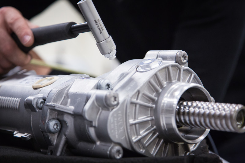 A close-up shot shows a hand holding a specialized Brüel & Kjær impact hammer, tapping the casing of a metallic automotive assembly. The mechanism features a prominent corrugated or threaded cylindrical shaft protruding from its side. The focus is sharp on the instrument tip and the immediate metal surface, while the person performing the test is softly blurred in the background.