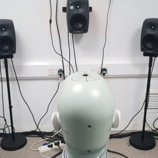 Copilot said: Head-shaped binaural measurement device surrounded by loudspeakers in an acoustic testing laboratory.