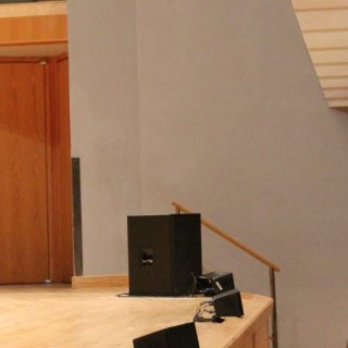 Presenter holding a saxophone on a concert hall stage while demonstrating acoustics to an audience.