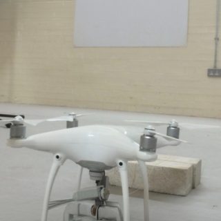 White quadcopter drone positioned in a large indoor testing facility with acoustic measurement equipment nearby.