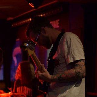 Musician playing electric guitar onstage under red lighting, with drummer blurred in the background.