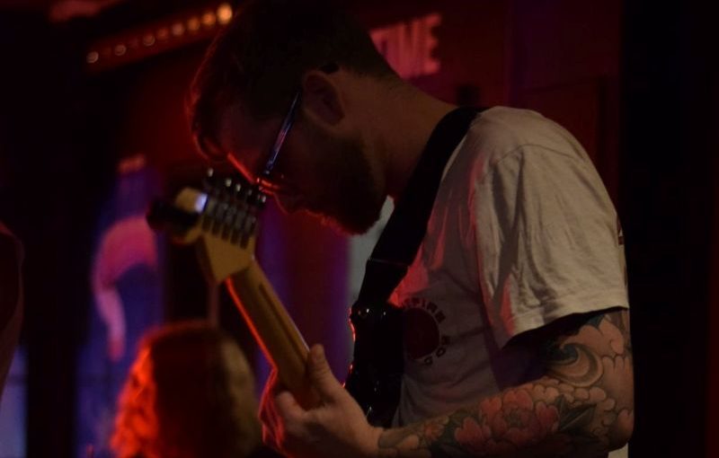 Musician playing electric guitar onstage under red lighting, with drummer blurred in the background.