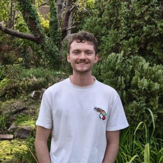 Person standing outdoors in a garden, wearing a white T-shirt, with trees and shrubs behind.