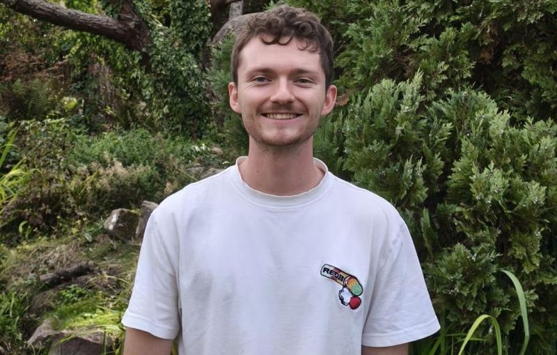 Person standing outdoors in a garden, wearing a white T-shirt, with trees and shrubs behind.